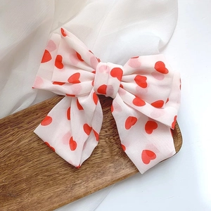 Detail of a large white chiffon bow hair clip featuring vibrant red heart patterns
