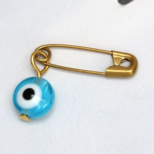 Detalle de un broche de imperdible dorado con cuenta de ojo turco azul sobre tela blanca