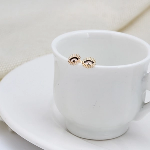Gold Evil Eye stud earrings with blue and black enamel displayed on a white ceramic cup