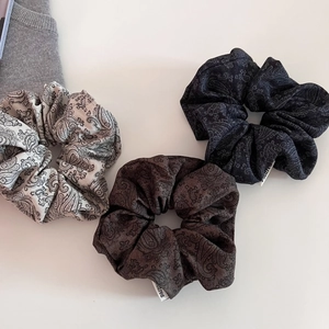 Close-up of three paisley pattern scrunchies in grey, brown, and navy on a white surface