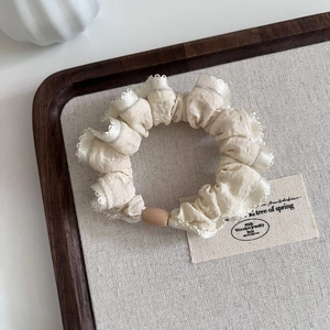 Elegant cream ruffled scrunchie with white lace trim and oval charm on a wooden tray