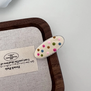 Close-up of an oval cream hair clip with colorful rainbow polka dots on a wooden tray