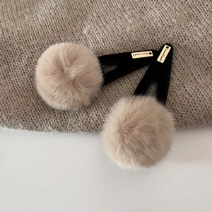 Detailed close-up of plush pom-pom hair clip featuring a golden handmade metal label tag