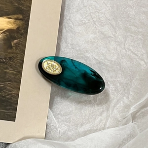 Detail of a dark green acetate oval hair clip with gold metal emblem on textured paper