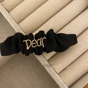 Close-up of black pleated fabric hair scrunchie with golden Dear metal lettering detail