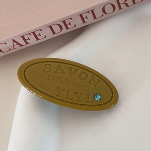 Detailed close-up of mustard yellow oval hair clip with embossed text and tiny gemstone