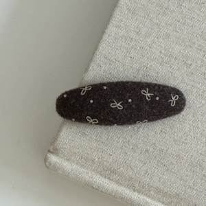 Dark brown felt oval hair clip with embroidered cream bow and dots on fabric background