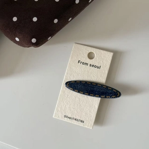 Oval denim fabric hair claw clip in dark blue with yellow stitching on display card