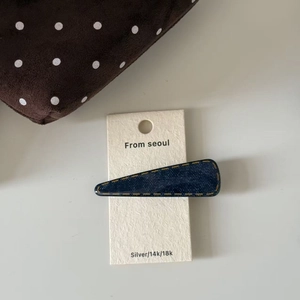 Detail of dark blue denim hair clip with yellow stitching on beige display card