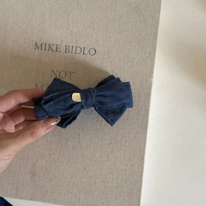 Detail view of dark blue denim bow hair clip with gold metal plaque held by hand