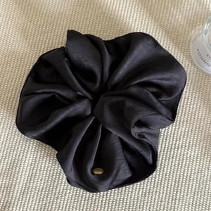 Detailed macro view of black satin hair scrunchie with golden metal logo charm and soft pleats