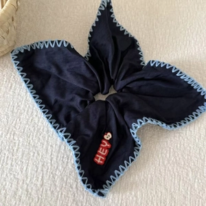 Detail of navy blue hair scrunchie with blue stitched edges and red HEY apple embroidery