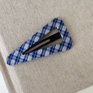 Detail showing a blue and white plaid fabric triangle hair clip on textured beige fabric