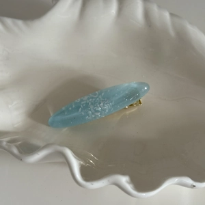 Barrette ovale en résine bleue avec flocons blancs et base en métal doré sur plateau coquillage
