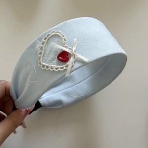 Detail eines blauen Spitzenstirnbandes mit weißem Herzflicken und rotem Knopf
