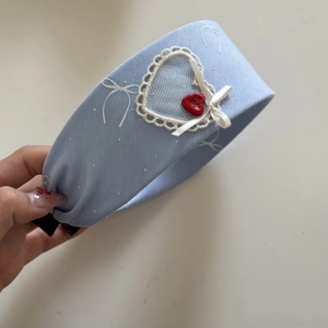 Detail eines hellblauen Spitzen-Herz-Stirnbands mit rotem Herzknopf und weißer Schleife