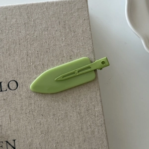 Matte avocado green flat alligator hair clip on a textured beige woven background