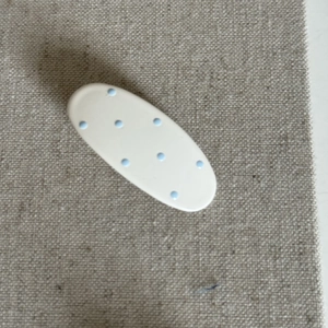 Close-up of cream-colored oval hair clip with light blue polka dots on a textured beige fabric