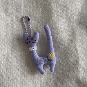 Detail of purple knitted cat plush keychain with pearl necklace and flower embroidery