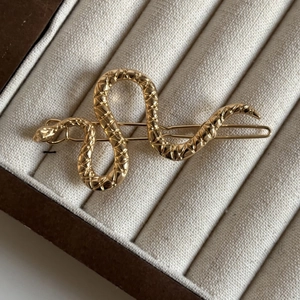 Luxurious gold snake-shaped metal hair clip with textured details on a beige display tray