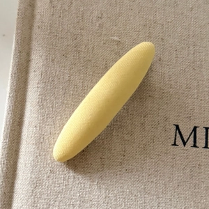 Cream yellow fabric-covered drop hair clip with minimalist oval shape on textured cloth