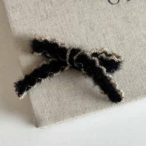 Elegant black plush bow hair clip with gold lace trim shown on a beige linen board
