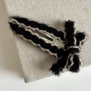 Detail of a plush black bow hair clip with beige trim and silver metal BB fastener