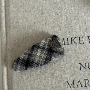 Grey plaid fabric hair clip with a vintage square metal badge on a textured background