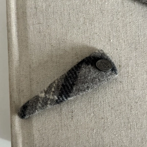 Detail view of a gray plaid wool hair clip with a vintage metal logo on linen background