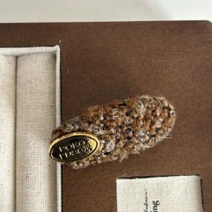 Brown wool tweed hair clip with gold metal badge shown on an elegant jewelry box display