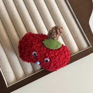 Handmade knitted red apple hair clip with cute cartoon eyes and plush texture on a tray