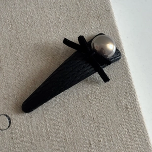 Detail of a black leather bow hair clip with a metallic grey pearl on textured fabric