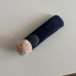 Detail of a blue knitted hair clip with a grey corduroy button and pink pom-pom decoration
