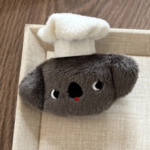 Close-up of a plush koala charm with a white chef hat on a beige linen display tray