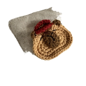 Handgefertigte gestrickte Capybara-Haarklammer mit rotem Hut auf grauem Sackleinen