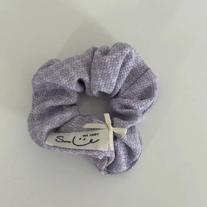 Detail eines lila Tweed-Haargummis mit cremefarbener Schleife und Webetikett