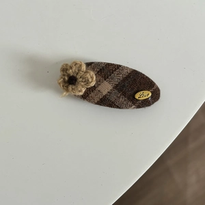 Detail of a brown plaid fabric hair clip with a knit flower and gold 'Love' metal tag