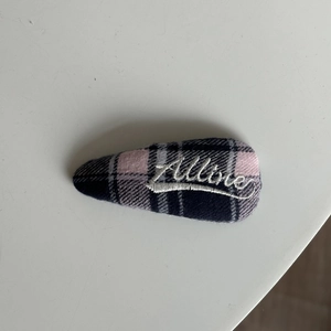 Dettaglio del fermaglio BB a quadri rosa e grigi con ricamo Allure in bianco
