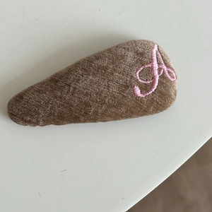 Detail of a brown corduroy hair clip featuring pink letter A embroidery on white background