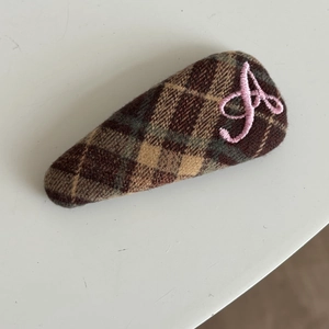 Detail of triangular hair clip featuring brown plaid fabric and pink letter A embroidery