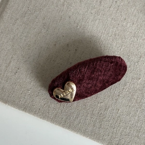 Detail view of burgundy velvet hair clip with golden heart ornament on beige fabric