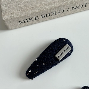 Detailed close-up of a blue knitted hair clip with silver sequins and metallic logo plaque