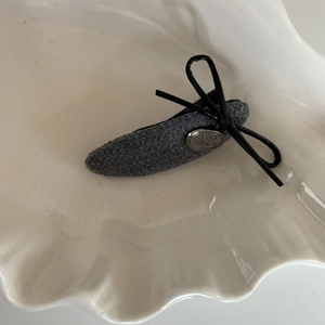 Elegant grey fabric hair clip with a black leather bow and silver metal logo on a tray