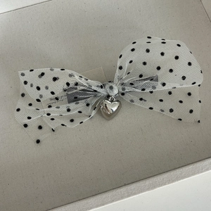Detail view of a white mesh bow hair clip with black polka dots and a silver heart charm