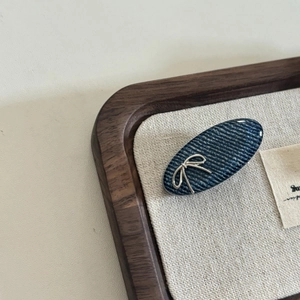 Denim blue oval hair clip with silver bow detail shown on a textured display tray
