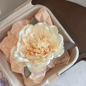 Detail view of a cream faux peony flower with green leaves in a kraft paper gift box