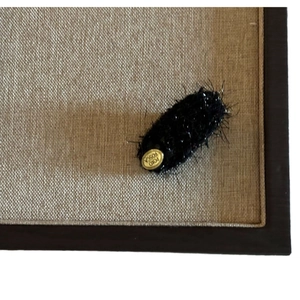 Plush brown oval hair clip with gold logo tag on a neutral woven textured background