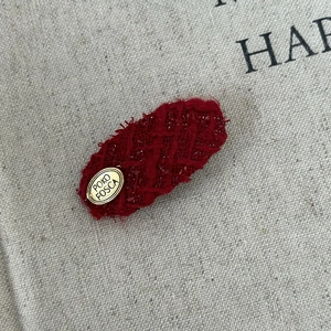 Red tweed hair clip with vintage golden logo plate on textured canvas background