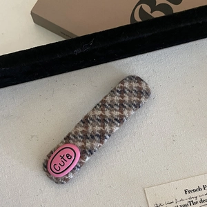 Detail of a brown plaid hair clip featuring a pink Cute label on textured fabric