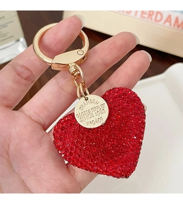 Detail showing hand holding a red rhinestone heart keychain with gold D-ring and engraved tag